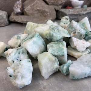 May include: A pile of raw, uncut gemstones. The stones are primarily white with patches and veins of light green and blue-green. The rough texture of the stones is visible, and they vary in size and shape. The background includes rocks and other decorative items.
