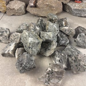 May include: A pile of rough-cut, gray and white granite rocks. The rocks vary in size and shape, with visible crystalline structures and dark flecks. The stones are arranged on a light-colored, tiled surface, suggesting a natural stone product.