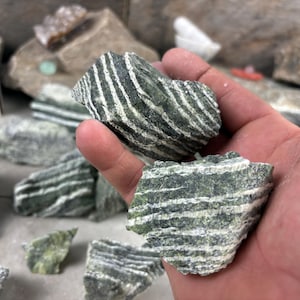 May include: Two pieces of green and white striped stone are held in a hand. The stones have a layered appearance, with alternating bands of green and white. Other similar stones are scattered in the background.
