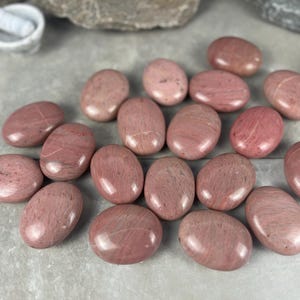 May include: A collection of polished, oval-shaped pink rhodonite stones. The stones vary in shade from light to deep pink, with some showing darker veining. The smooth, glossy surfaces reflect light, highlighting their natural beauty. A mortar and pestle are in the background.
