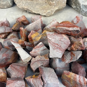 May include: A pile of rough-cut rocks in shades of red, gray, and yellow. The stones have a layered appearance with visible mineral striations. The rocks vary in size and shape, with some appearing angular and others more rounded.