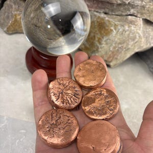 May include: Five circular copper discs with textured, uneven surfaces resting in a palm. The discs have a metallic, reddish-brown finish with varying degrees of surface detail, ranging from smooth to cracked patterns, displayed alongside a clear glass sphere.
