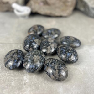 May include: A collection of tumbled stones with a speckled pattern. The stones are oval-shaped and feature a mix of gray, blue, and white colors. They are arranged on a light gray surface.