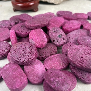 May include: A collection of numerous small, oval-shaped, vibrant magenta-colored stones with a porous, textured surface. Each stone features a flat, disc-like profile with visible indentations and a rough, natural finish across the entire surface.