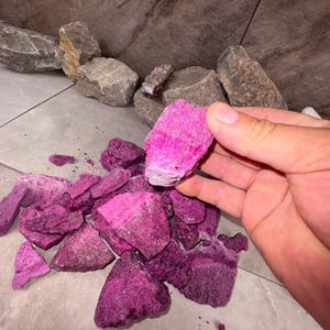 May include: A collection of vibrant, magenta-colored rocks and stones. The rocks vary in size and shape, with a hand holding one of the larger pieces. The background features larger gray and brown stones, and a light-colored tiled surface.