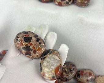 Puede incluir: Un conjunto de piedras pulidas de forma ovalada con apariencia de mosaico. Las piedras presentan una mezcla de colores rojo, negro, marrón y beige. Algunas piedras se sostienen en una mano enguantada, mientras que otras están sobre una superficie blanca.