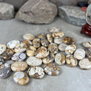 May include: A collection of polished, oval-shaped stones in shades of cream, brown, and gray. The stones have unique patterns and textures, with a few larger rocks and a crystal ball in the background.