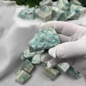 May include: A hand wearing a white glove holds a piece of aquamarine gemstone. The stone is a mix of light blue and white with darker blue and silver flecks. Other similar stones are scattered in the background on a white surface.