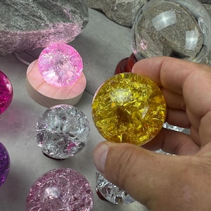 May include: A collection of decorative glass spheres in various colors, including pink, purple, yellow, and clear. One sphere is held in a hand. Some spheres are illuminated, and one rests on a wooden base. The spheres have a cracked, textured appearance.