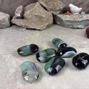 May include: A collection of polished agate stones in shades of green, black, and white. The stones have a smooth, rounded shape and display unique banding patterns. The arrangement is on a neutral surface, with rocks and a crystal ball in the background.