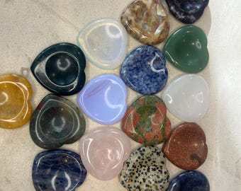 May include: Assortment of heart-shaped polished stones in various colors and patterns, including blue, green, pink, red, brown, and white. The stones are arranged in a semi-circle on a white surface.