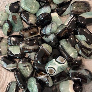 May include: A collection of polished agate stones in shades of black, white, and green. The stones vary in size and shape, with some displaying banded patterns and others featuring circular designs. The gemstones are smooth and glossy.