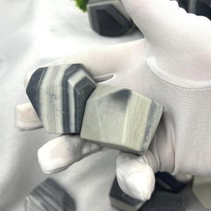 May include: Geometric-shaped stones with gray and white striped patterns. The stones are held in a white gloved hand, with several more stones visible in the background. The stones have a polished appearance and a natural aesthetic.