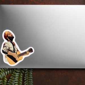 K&ouml;nnte beinhalten: Ein Aufkleber mit einer Illustration einer Person, die eine Akustikgitarre spielt. Die Person tr&auml;gt einen Turban und ein gemustertes Hemd. Der Aufkleber befindet sich auf einem silbernen Laptop, mit einem Farn und einem dunkelbraunen Hintergrund.