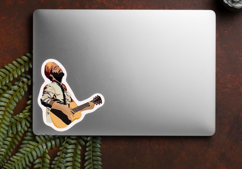 K&ouml;nnte beinhalten: Ein Aufkleber mit einer Illustration einer Person, die eine Akustikgitarre spielt. Die Person tr&auml;gt einen Turban und ein gemustertes Hemd. Der Aufkleber befindet sich auf einem silbernen Laptop, mit einem Farn und einem dunkelbraunen Hintergrund.