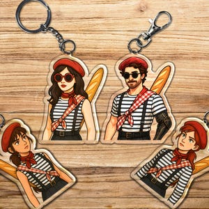 Puede incluir: Llaveros con ilustraciones de dibujos animados de personas con atuendo francés, incluyendo boinas, camisas a rayas y bufandas rojas. Cada llavero tiene una baguette y está unido a un llavero plateado.