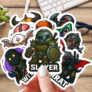 Può includere: Una collezione di adesivi colorati a cartoni animati con vari personaggi fantasy. Gli adesivi includono una figura verde corazzata con uno scudo, un teschio e altre figure mostruose. La parola "SLAYER" è visibile su un adesivo.