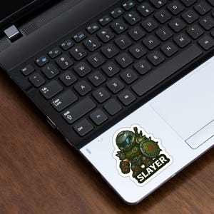 Pode incluir: Um autocolante com uma ilustra&ccedil;&atilde;o de desenho animado de um personagem com armadura verde, segurando um escudo. A palavra "SLAYER" est&aacute; escrita em branco sobre um ret&acirc;ngulo preto. O autocolante est&aacute; no touchpad de um laptop.