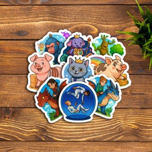 以下が含まれることがあります： 様々な漫画のキャラクターとファンタジー要素が特徴の、カラフルなステッカーのコレクション。豚、猫、象、ドラゴン、スノードームの中の人などのデザインが含まれています。ステッカーは木製の表面に配置されています。