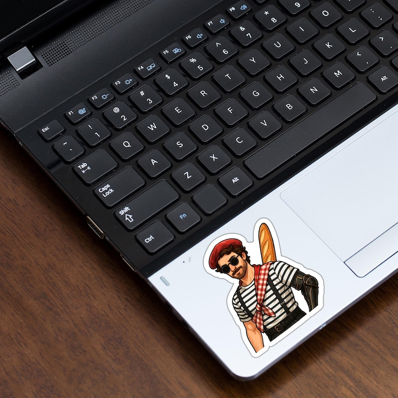 May include: A sticker depicting a cartoon man in a striped top, red beret, and sunglasses, holding a baguette. The sticker is on a laptop's trackpad, with the keyboard in view.
