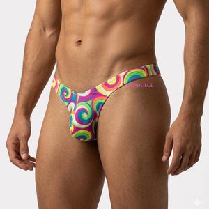 May include: A colorful thong swimsuit featuring a vibrant swirl pattern in shades of pink, green, yellow, and purple. The swimsuit has a low-rise design and a narrow waistband. The word "ANDULCE" is printed on the side.
