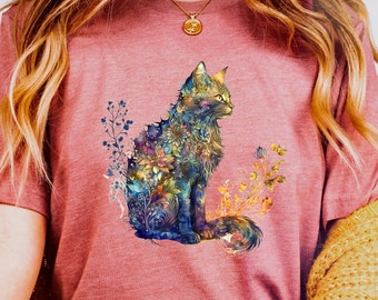 May include: A woman wearing a maroon t-shirt with a floral cat design. The cat is outlined in black and filled with colorful flowers. The woman is wearing blue jeans and a yellow cardigan.