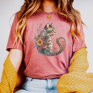 May include: A woman wearing a pink t-shirt with a floral design featuring a cat. The cat is surrounded by flowers and leaves in shades of blue, green, purple, and yellow. The woman is also wearing blue jeans and a yellow cardigan.