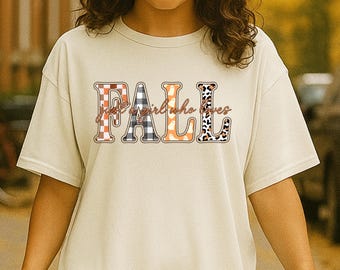 May include: Cream-colored t-shirt with the word "FALL" in a mix of plaid, orange, and leopard print patterns. The text "a girl who loves" is above the word "FALL". The person is holding a coffee cup.