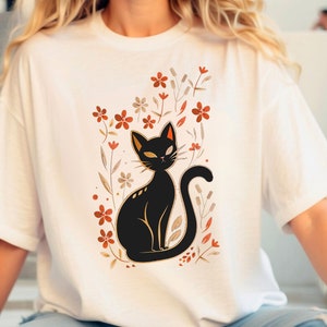 May include: White t-shirt featuring a black cat illustration with orange and brown floral accents. The cat has yellow eyes and is surrounded by stylized flowers and foliage. The design is centered on the shirt.