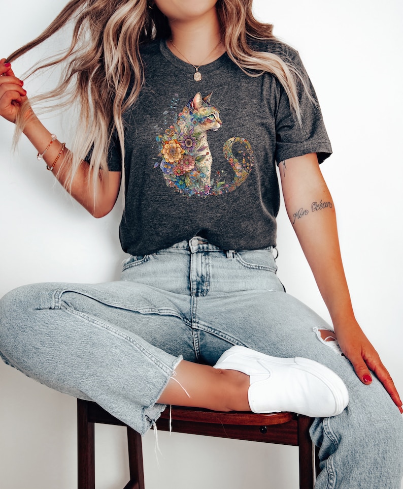 May include: A dark grey t-shirt featuring a colourful floral cat design. The cat is looking to the right and has a light brown and white coat. The flowers are in shades of pink, yellow, orange, and green.