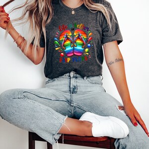 May include: Dark gray t-shirt featuring a colorful graphic of rainbow-colored flip-flops. The text "Life is better in Flip Flops" is printed above and below the flip-flops. The shirt is made of a soft material.