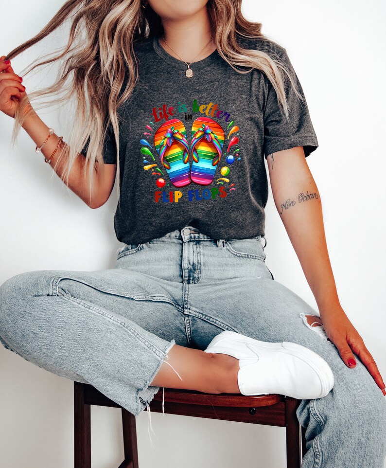 May include: Dark gray t-shirt featuring a colorful graphic of rainbow-colored flip-flops. The text "Life is better in Flip Flops" is printed above and below the flip-flops. The shirt is made of a soft material.