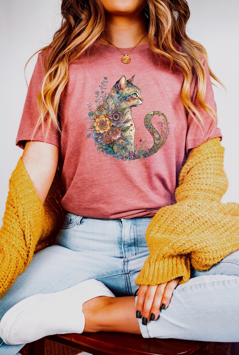 May include: A woman wearing a pink t-shirt with a floral design featuring a cat. The cat is surrounded by flowers and leaves in shades of blue, green, purple, and yellow. The woman is also wearing blue jeans and a yellow cardigan.