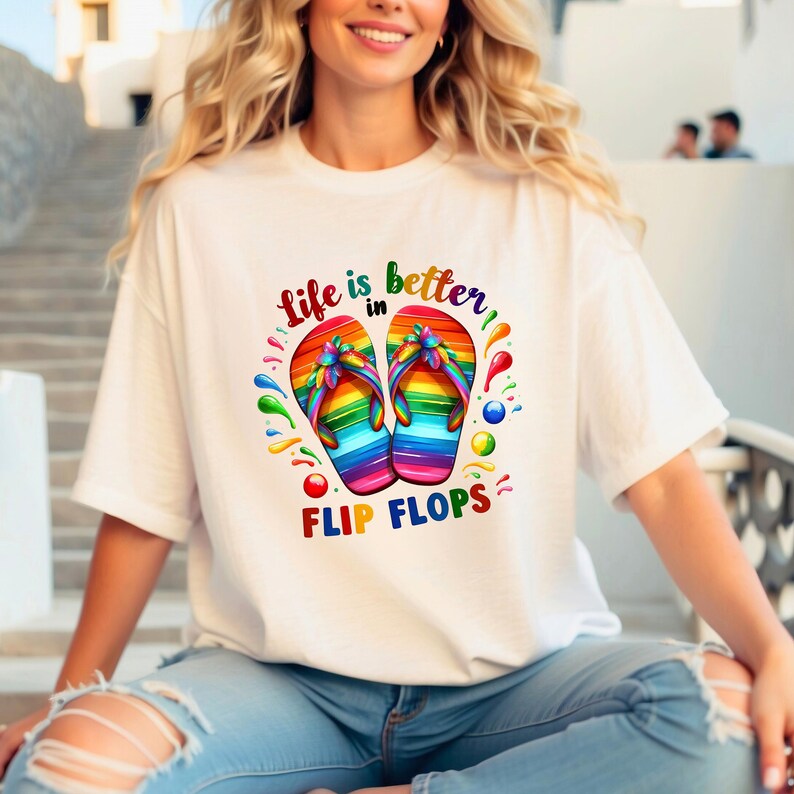 May include: White t-shirt with a colorful graphic that reads "Life is better in Flip Flops." The graphic features two rainbow-colored flip-flops and colorful splashes. The shirt is a casual, relaxed fit.