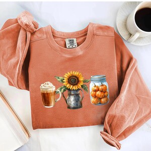 Könnte beinhalten: Ein orangefarbenes Langarmshirt mit einem herbstlichen Motiv. Das Motiv zeigt eine Sonnenblume in einem Metallkrug, einen Latte und ein Glas voller Kürbisse. Eine weiße Kaffeetasse befindet sich in der oberen rechten Ecke.