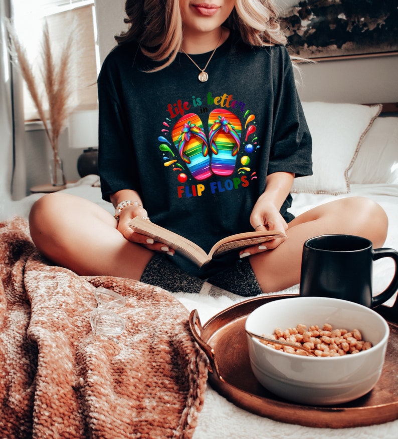 May include: Dark gray t-shirt with a colorful graphic of rainbow flip-flops and the text "Life is better in Flip Flops." The shirt is worn by a person sitting on a bed with a book, a mug, and a bowl of cereal on a tray.