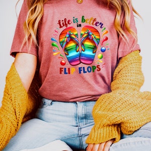 May include: A maroon t-shirt with the text "Life is better in Flip Flops." The shirt features a colorful graphic of rainbow-colored flip-flops. The shirt is paired with light blue jeans and a yellow sweater.