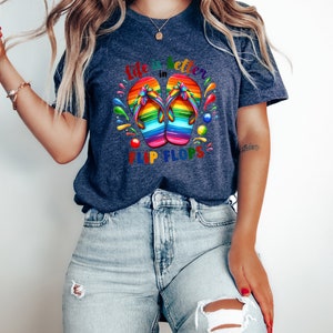 May include: Navy blue t-shirt with the text "Life is better in Flip Flops." The shirt features a colorful graphic of rainbow-colored flip-flops. The design includes splashes of color around the flip-flops, creating a vibrant and summery feel.