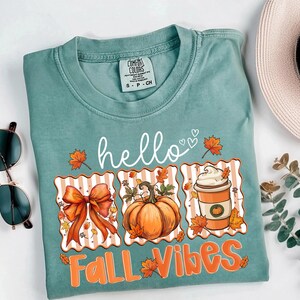 以下が含まれることがあります： 「hello FALL VIBES」の文字が白とオレンジでプリントされた、ティール色のComfort Colors Tシャツ。リボン、カボチャ、葉っぱのコーヒーカップなど、秋をテーマにしたグラフィックが特徴です。