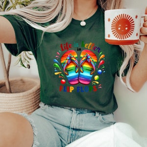 May include: Dark green t-shirt with the text "life is better in flip flops" and a graphic of rainbow-colored flip-flops. A white mug with a sun design is held in the hand.