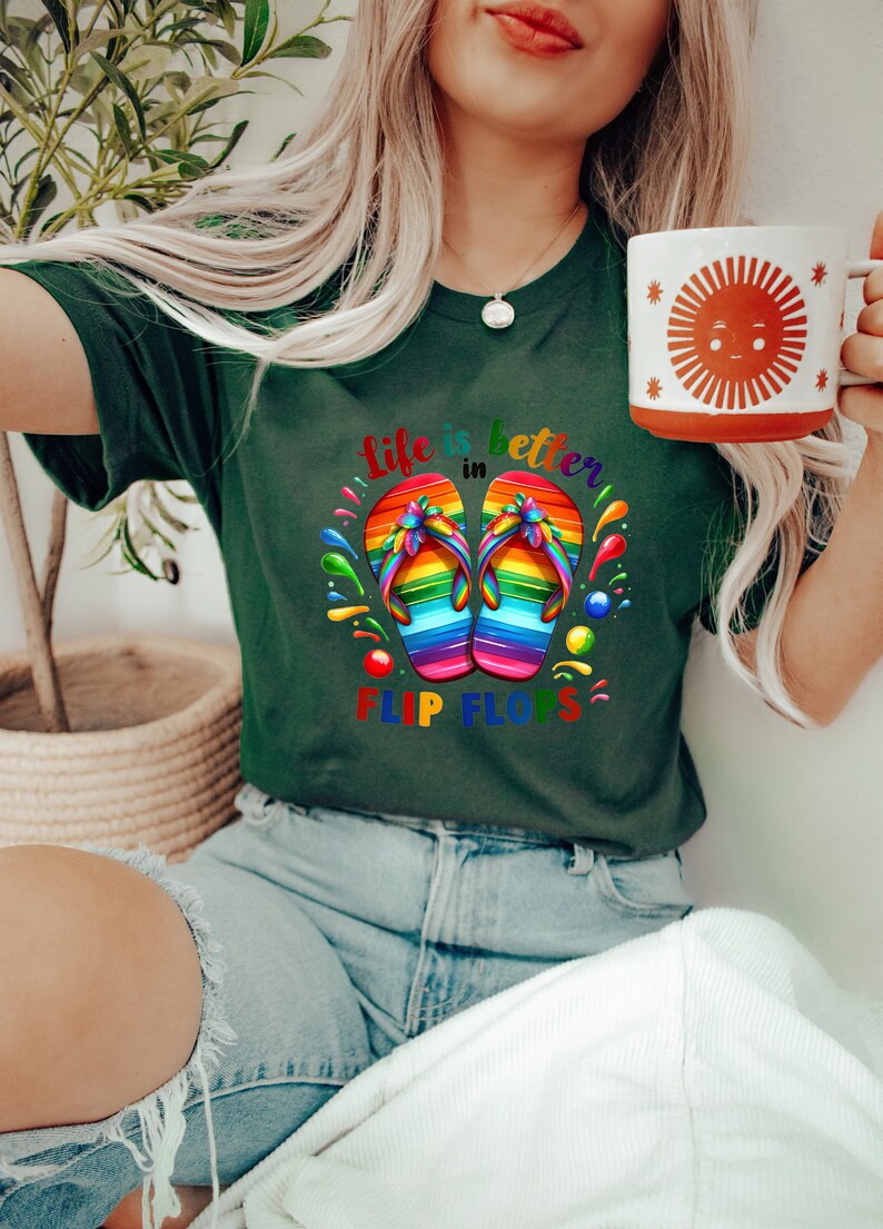 May include: Dark green t-shirt with the text "life is better in flip flops" and a graphic of rainbow-colored flip-flops. A white mug with a sun design is held in the hand.