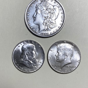 May include: Three silver coins: a Morgan silver dollar from 1884, a 1953 Franklin half dollar, and a 1966 Kennedy half dollar. The coins are displayed on a light gray surface.