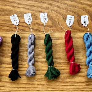 May include: A collection of colorful embroidery floss skeins, each with a small tag. The skeins are in various colors, including white, purple, black, green, red, blue, and peach. The floss is arranged on a wooden surface.