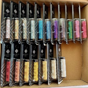 May include: A variety of colorful spools of thread in clear plastic packaging, arranged in a cardboard box. The threads are in shades of pink, gold, blue, purple, and green. Each spool is labeled with Japanese text.