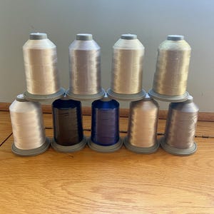 Puede incluir: Un surtido de diez carretes de hilo de bordar en varios colores metálicos y lisos. Los colores incluyen plata, oro, negro, azul y bronce. Los carretes están dispuestos en dos filas, con cuatro carretes en la fila superior y seis en la inferior.