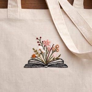 Op de afbeelding: Crèmekleurige draagtas met geborduurd bloemmotief. De borduursels tonen een open boek met kleurrijke bloemen, waaronder roze, oranje en witte bloesems. De tas heeft twee lange handvatten.