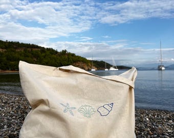 May include: A beige tote bag with embroidered sea-themed designs, including a starfish, seashell, and spiral shell. The bag is on a pebble beach with a scenic ocean and sky background.