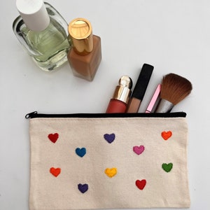 May include: A beige canvas makeup bag with a black zip, embroidered with colourful hearts. The bag is open, revealing makeup brushes and bottles. A perfume bottle and foundation bottle are also visible.
