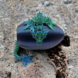 May include: A 3D printed dragon figurine with a green and blue color scheme. The dragon is curled up on a black egg-shaped base.