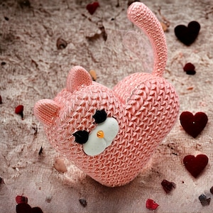 May include: A pink crocheted heart-shaped cat figurine with white eyes and a white nose. The figurine is surrounded by red hearts on a brown textured background.